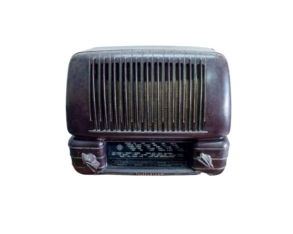 radio de mesa de la prestigiosa marca Telefunken, caracterizada por su robusto cuerpo de baquelita en tono oscuro con líneas redondeadas, una rejilla frontal con barras verticales y un panel inferior de diales con inscripciones detalladas de emisoras y frecuencias alquiler para eventos, publicidad, peliculas y series