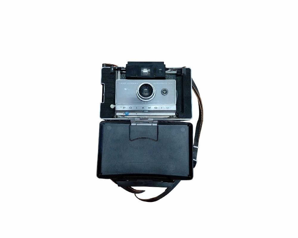 cámara fotográfica instantánea clásica de fuelle de la reconocida marca Polaroid, modelo Automatic 104 Land Camera, que se muestra abierta con su cuerpo metálico y plástico, lente central y tapa abatible inferior junto con su correa de transporte alquiler para eventos, publicidad, peliculas y series