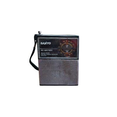 radio portátil de época de la marca Sanyo, caracterizada por su carcasa rectangular compacta con asa lateral, un panel superior oscuro que incluye un dial de sintonía analógico de forma circular y una rejilla metálica frontal perforada inferior alquiler para eventos, publicidad, peliculas y series