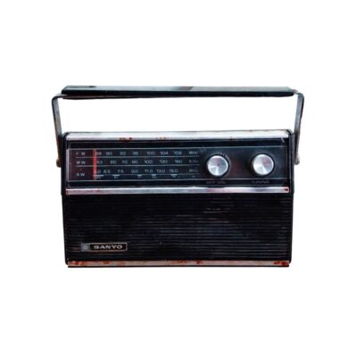 radio portátil de época de la marca Sanyo, caracterizada por su carcasa rectangular oscura con asa de transporte superior, un panel frontal superior que integra un amplio dial analógico multibanda y mandos circulares, y una rejilla inferior para el altavoz alquiler para eventos, publicidad, peliculas y series