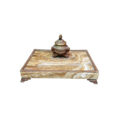 tintero de escritorio de época elaborado en piedra de ónix natural con vetas cálidas, caracterizado por una base rectangular con remates y patas metálicas cinceladas, y un tintero central de bronce con tapa y detalles ornamentales alquiler para eventos, publicidad, peliculas y series